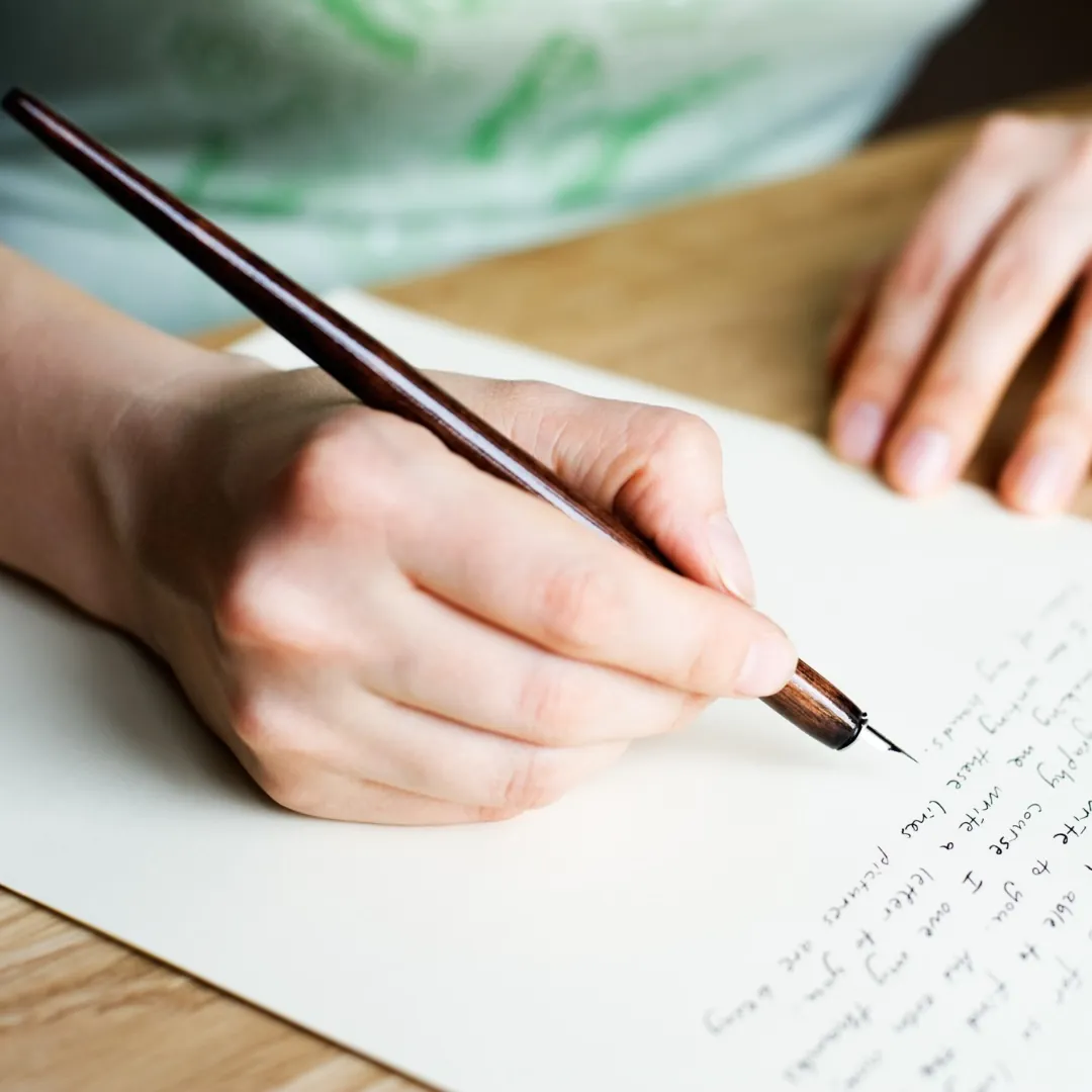 Close-up of a person’s hand writing on cream-colored paper with a dark wood fountain pen. The scene captures a moment of journaling or letter writing on a wooden desk.