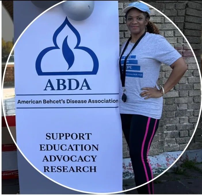 A woman standing next to a banner for the American Behcet’s Disease Association (ABDA). The banner features the ABDA logo and text that reads: 'American Behcet’s Disease Association' followed by 'Support, Education, Advocacy, Research.'