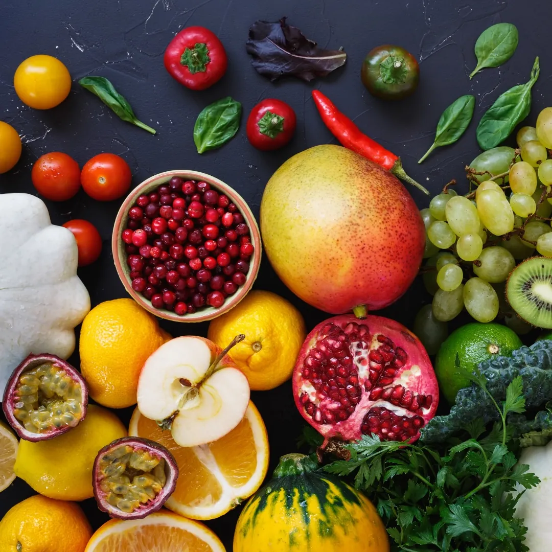 An assortment of colorful fresh fruits and vegetables arranged on a dark surface, including grapes, kiwi, mango, pomegranate, apple, oranges, passion fruit, cranberries, tomatoes, peppers, leafy greens, and herbs.