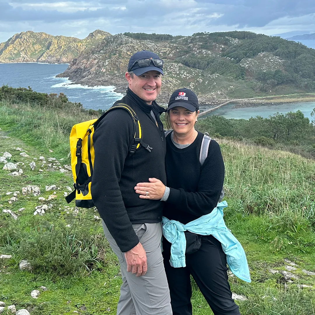 A smiling couple in hiking gear poses together on a scenic trail with rocky hills, lush greenery, and ocean waves in the background. The man wears a yellow backpack and sunglasses, while the woman has a light blue jacket tied around her waist. They appear happy and relaxed, enjoying the outdoor adventure.