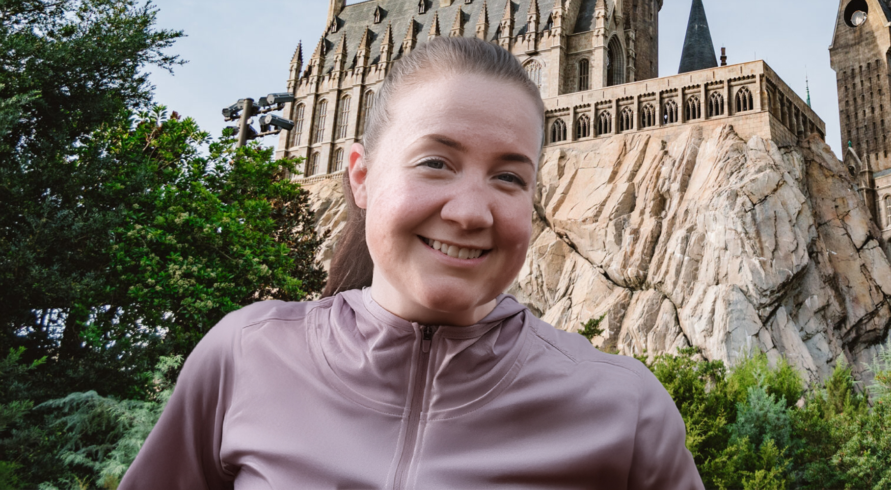 A woman smiling outdoors, wearing a light purple zip-up top. Behind her is a large stone castle-like building with tall windows and towers, built into a rocky hillside, surrounded by greenery.