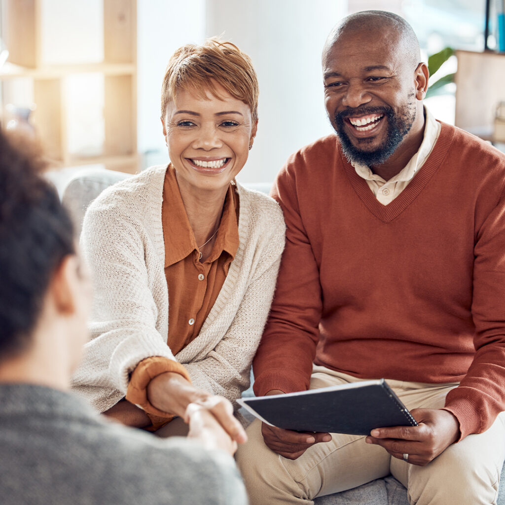 A smiling couple sits together, one holding a notebook, while shaking hands with another person in the foreground. The couple appears happy and engaged in discussion.
