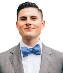 A man with short dark hair wearing a light gray suit, white shirt, and blue bow tie, smiling slightly against a white background.