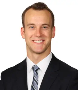 A man with short light brown hair wearing a black suit, white shirt, and striped tie, smiling broadly against a white background.