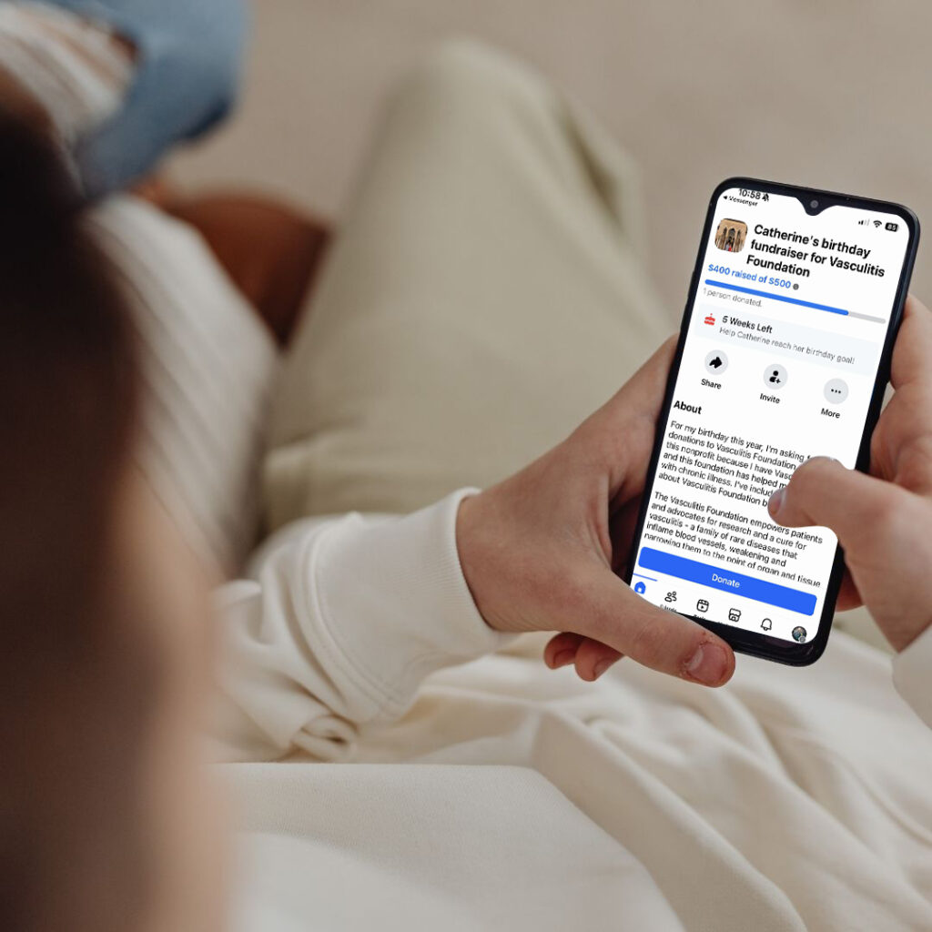 A person holding a smartphone while viewing a Facebook birthday fundraiser page for the Vasculitis Foundation. The screen shows the fundraiser progress bar, goal, and description text.