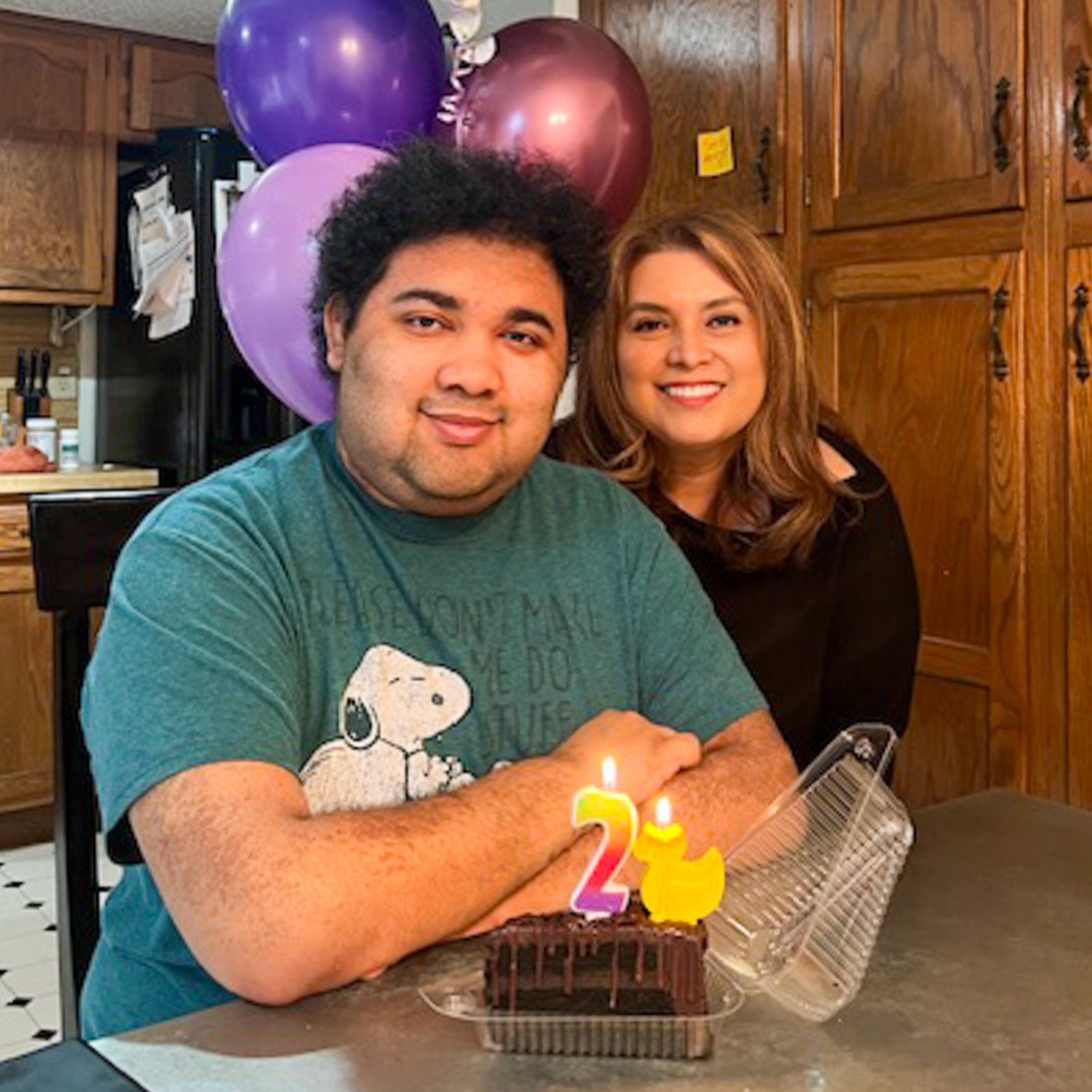 A young man in a teal Snoopy T-shirt sits at a table with a chocolate cake topped with candles shaped as the number 21. Behind him, a smiling woman with long brown hair leans close. Purple balloons and wooden cabinets are visible in the background.