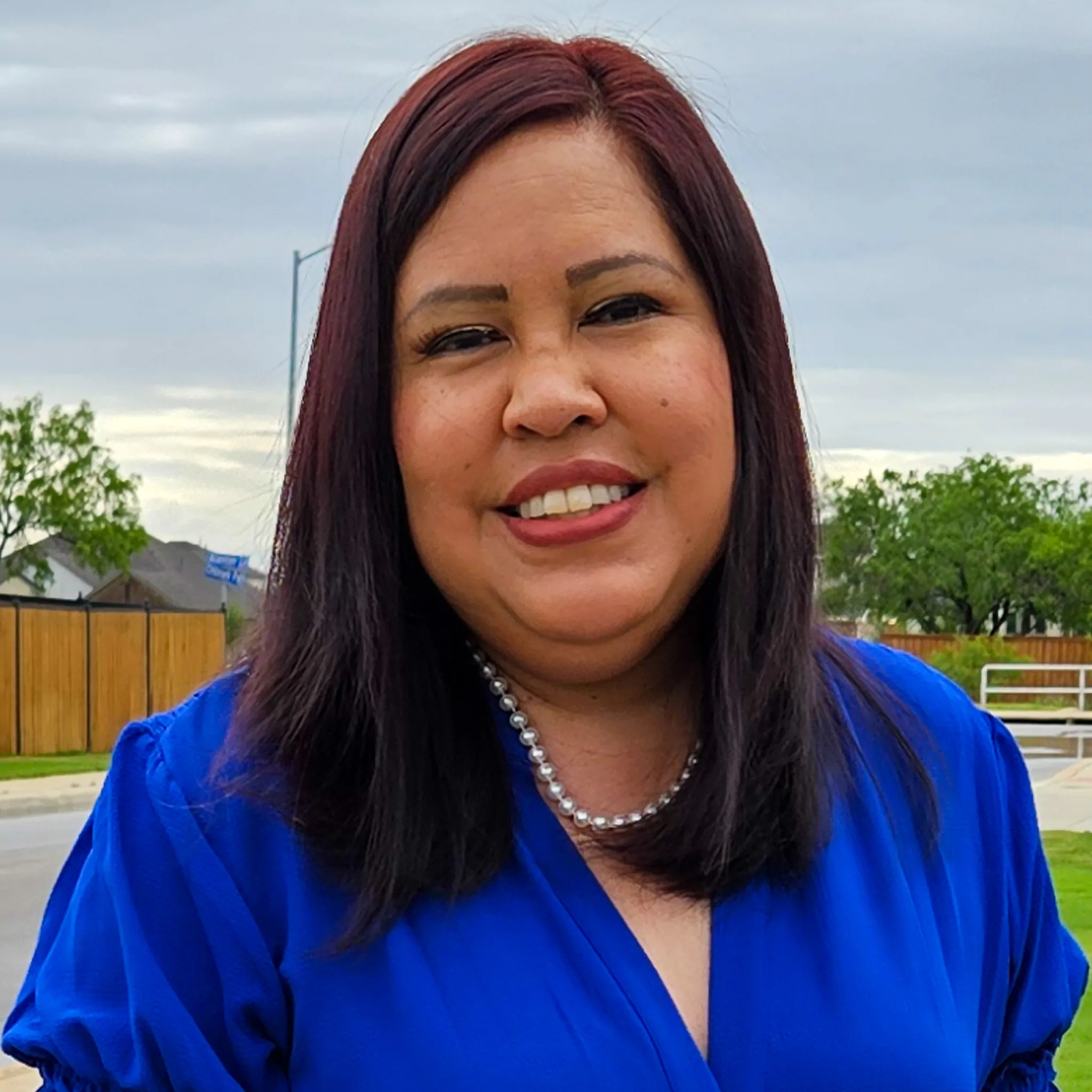 A woman with straight dark red hair, wearing a bright blue blouse and a pearl necklace, smiles outdoors. Trees, a fence, and a cloudy sky are visible in the background.