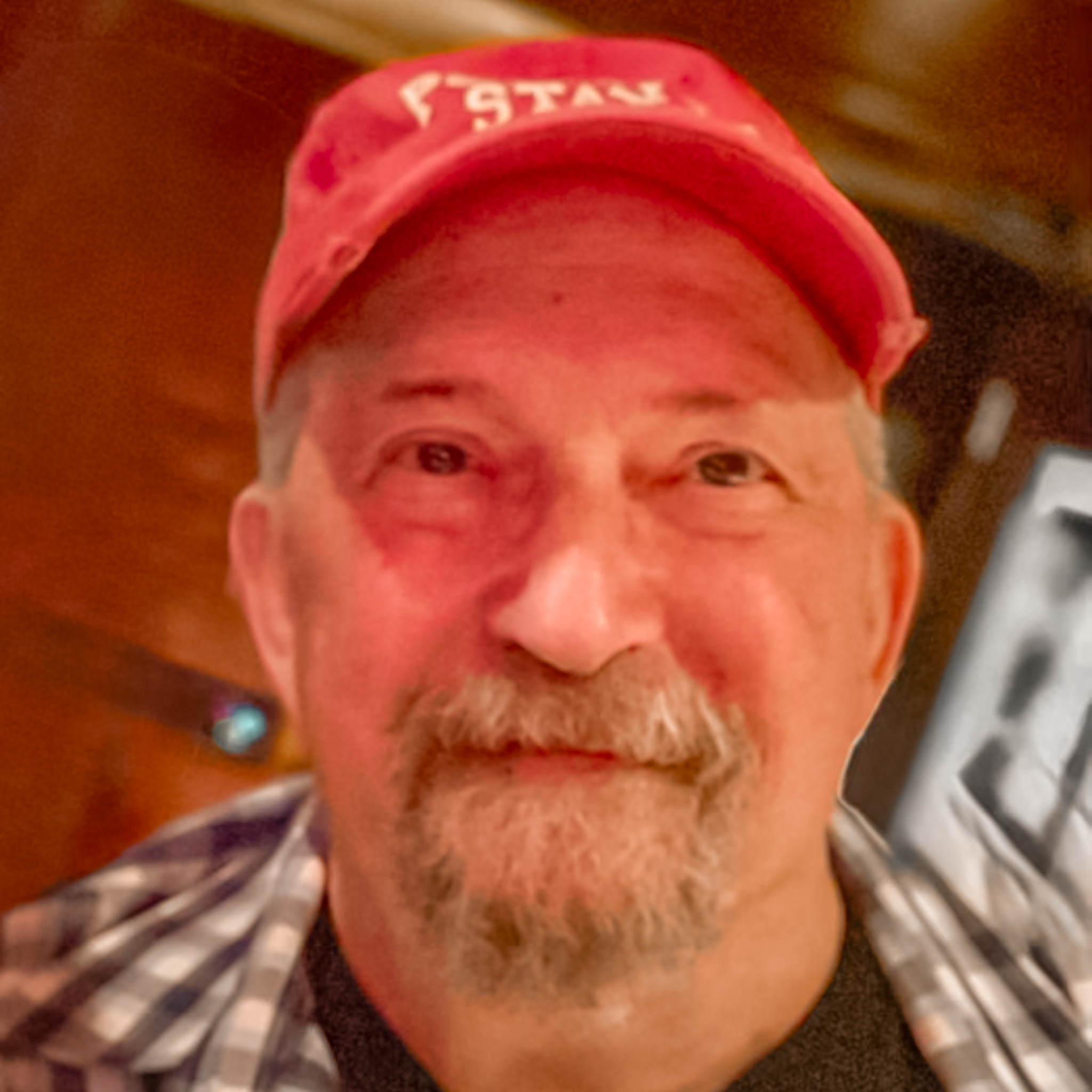 A man with a mustache and goatee wearing a red cap and a plaid shirt smiles at the camera indoors with a warmly lit background.
