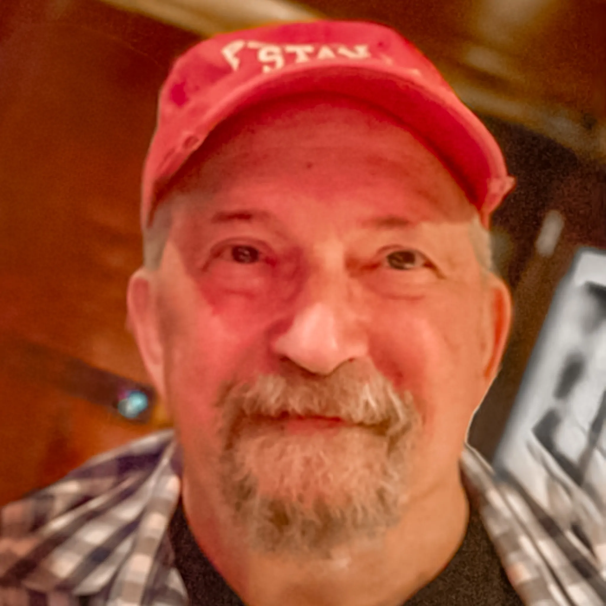 A man with a mustache and goatee wearing a red cap and a plaid shirt smiles at the camera indoors with a warmly lit background.