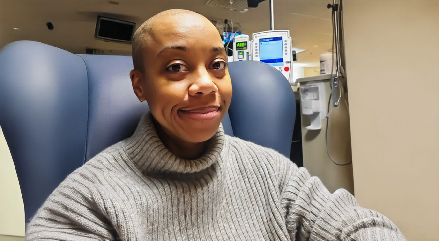 A woman with a shaved head sits in a medical chair, wearing a gray turtleneck sweater. She looks at the camera with a gentle smile. Medical equipment and monitors are visible in the background, suggesting a treatment setting.