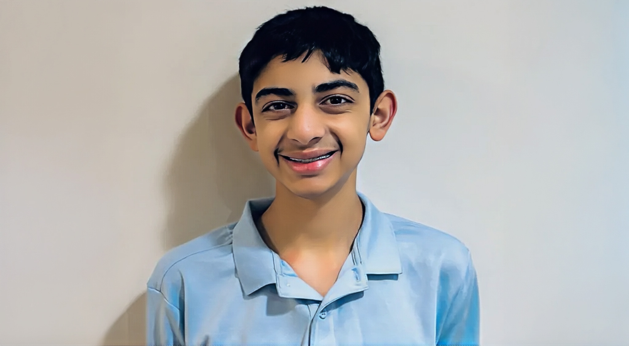 A smiling boy with short dark hair stands against a plain light-colored wall. He is wearing a light blue collared shirt.
