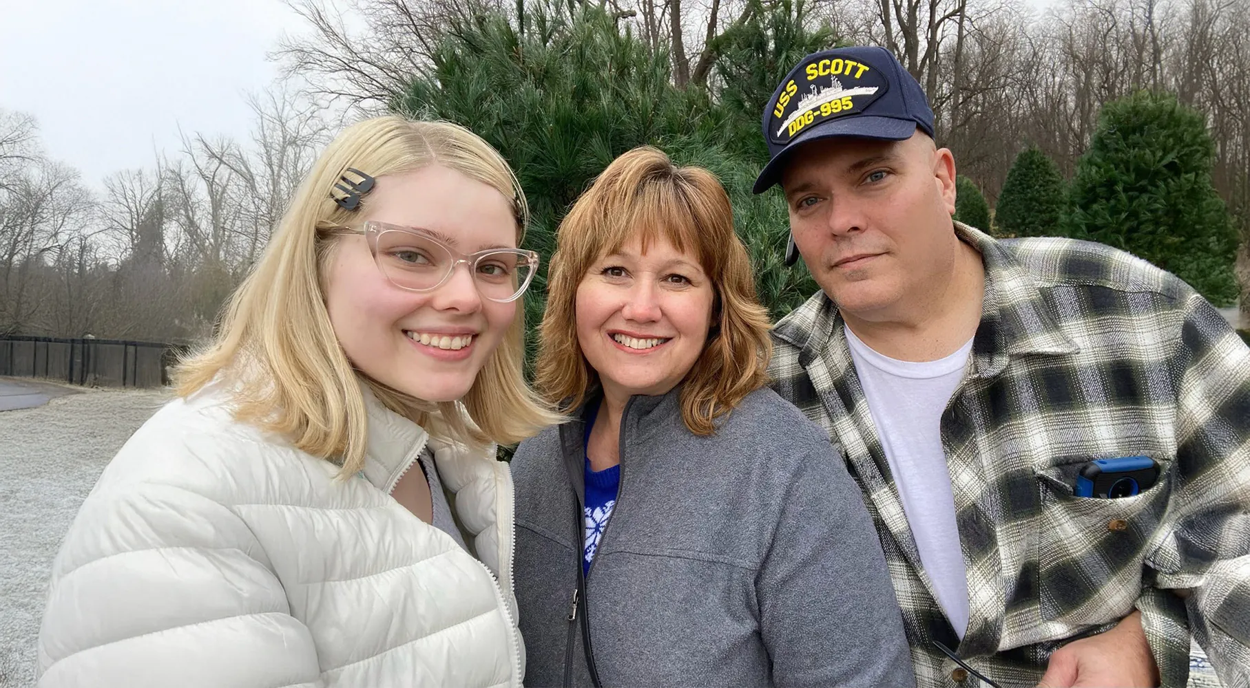 “Three people stand close together outdoors, smiling at the camera. A young woman in a white jacket is on the left, an adult woman in a gray jacket is in the center, and an adult man wearing a plaid shirt and a navy USS Scott cap is on the right. Evergreen trees and bare winter woods are in the background.