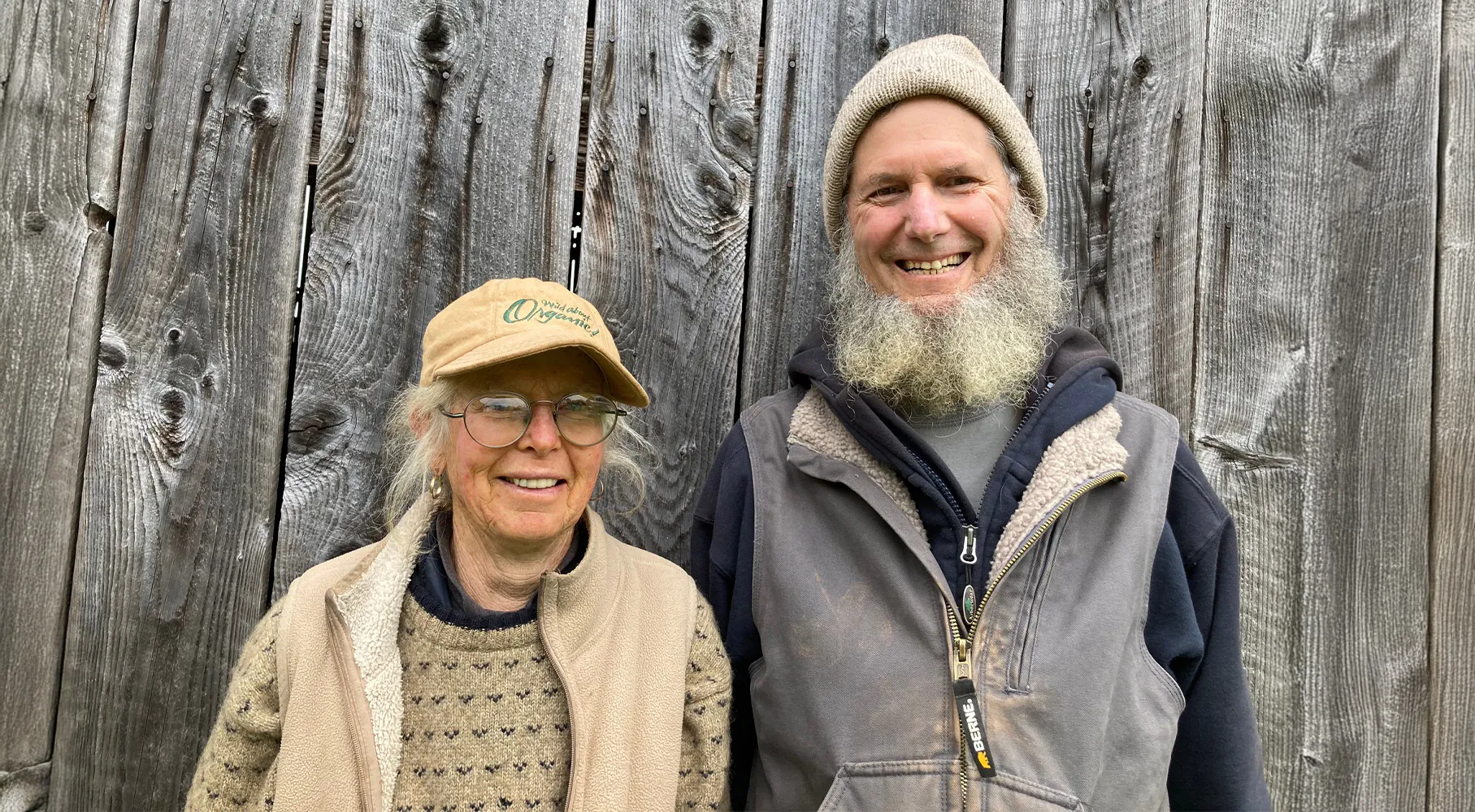Two older adults stand smiling in front of a weathered wooden wall. The woman on the left wears glasses, a tan cap, and layered outdoor clothing. The man on the right has a long gray beard and wears a knit hat and a zip-up vest over a hoodie.