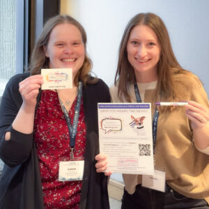 Two women smile while holding promotional materials for the VPPRN Pediatric Vasculitis Registry. The woman on the left holds a small VPPRN card, and the woman on the right holds a flyer and a purple pen. Both wear conference lanyards and stand together indoors.