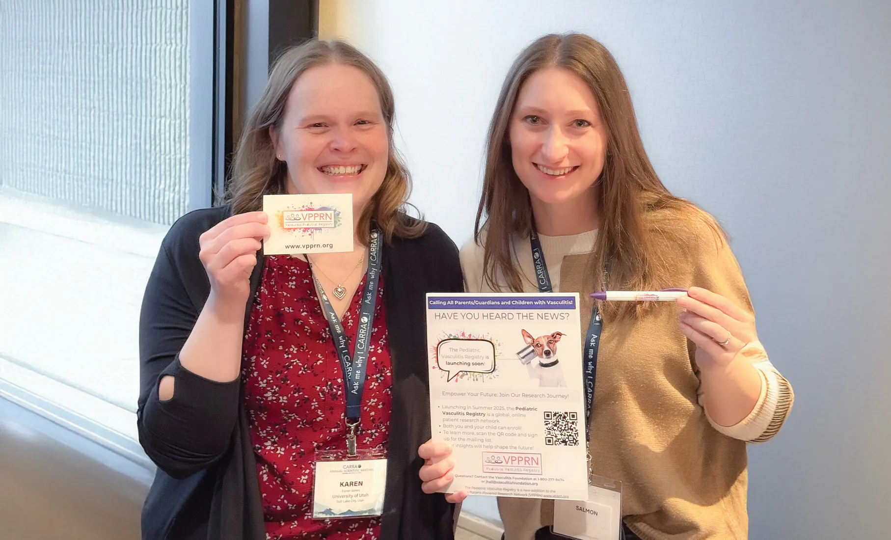 Two women smile while holding promotional materials for the VPPRN Pediatric Vasculitis Registry. The woman on the left holds a small VPPRN card, and the woman on the right holds a flyer and a purple pen. Both wear conference lanyards and stand together indoors.