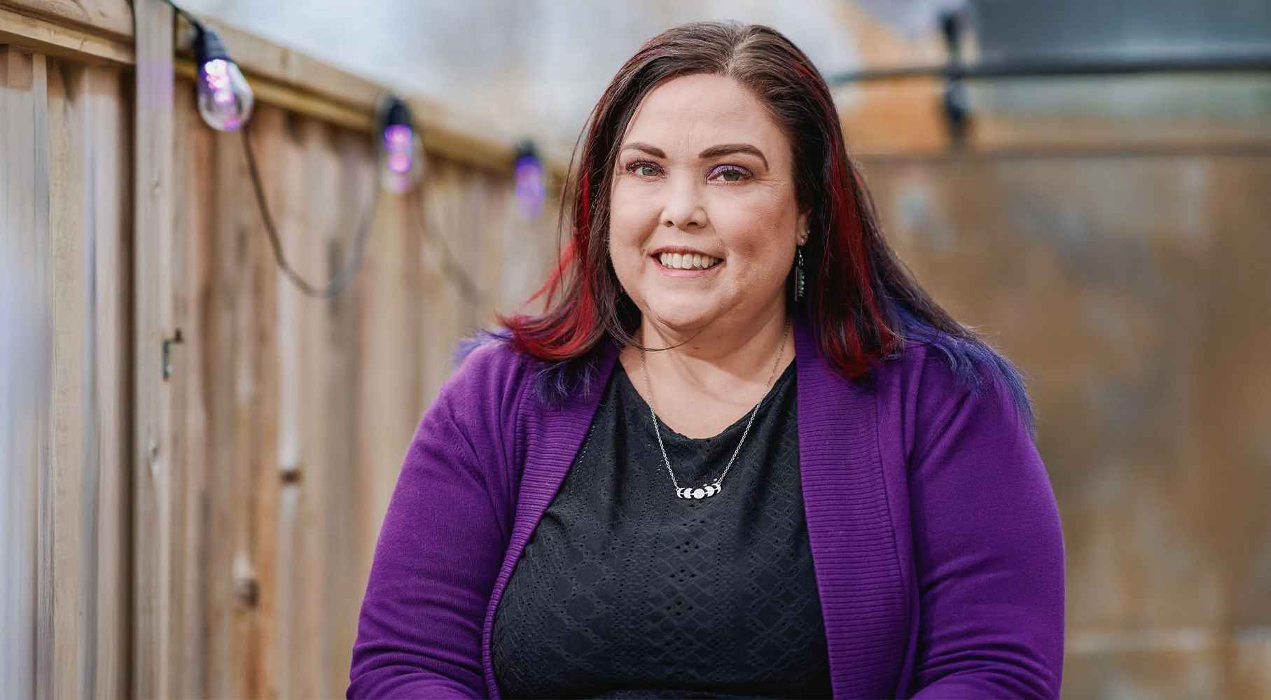 A woman with dark hair streaked with red sits outdoors, smiling at the camera. She is wearing a purple cardigan over a black top and a moon-phase necklace. A wooden fence with string lights hangs behind her
