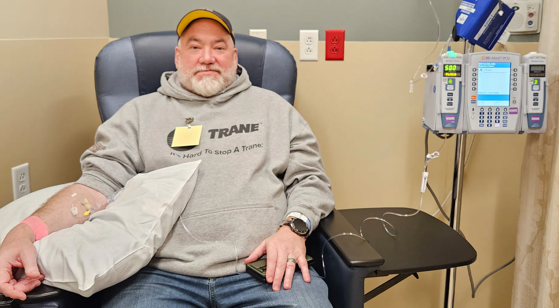 A man sits in a medical infusion chair with an IV in his arm, resting on a pillow. He wears a gray hoodie and a yellow baseball cap, looking calmly at the camera. An infusion pump stands to his right with tubes connected to his IV