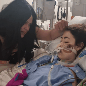 Imogen in hospital bed with family member at her bed side.