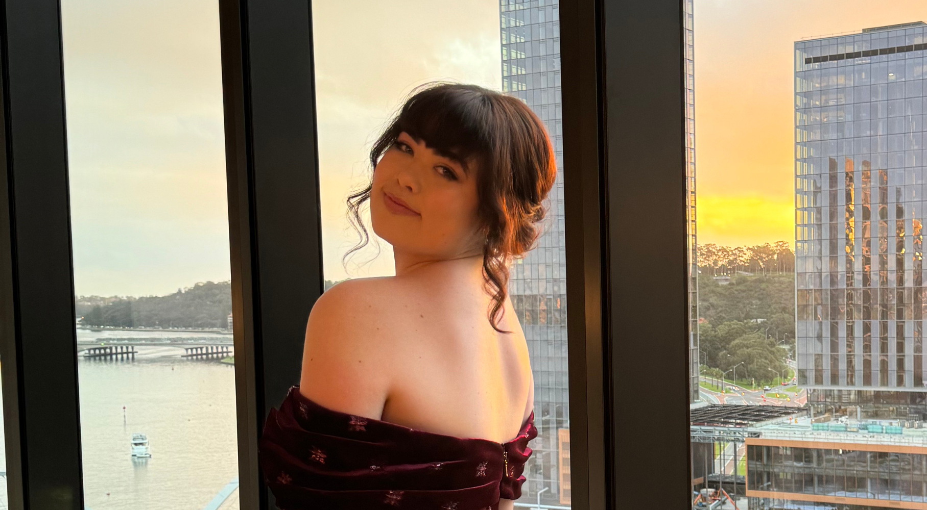 A woman in an off-the-shoulder burgundy dress stands by a tall glass window overlooking a waterfront at sunset. She is turned slightly toward the camera with a soft smile, with modern buildings and boats visible outside across the water.