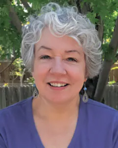 Smiling woman with short gray hair wearing a purple top and dangling earrings, photographed outdoors with greenery in the background.