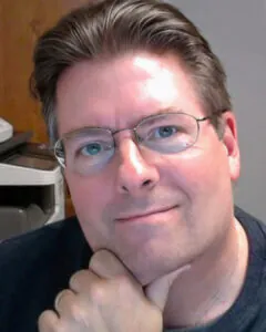 Man wearing glasses resting his chin on his hand, photographed indoors with office equipment visible in the background.