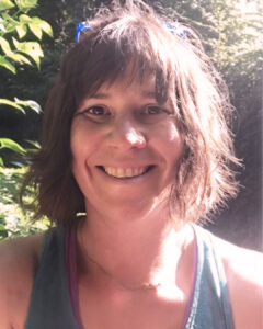 Smiling woman outdoors with short brown hair, sunlight filtering through trees behind her.