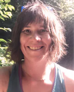 Smiling woman outdoors with short brown hair, sunlight filtering through trees behind her.