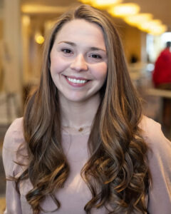Smiling woman with long brown hair in an indoor setting with warm lighting and blurred background.