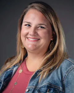 Smiling woman with long light brown hair wearing a denim jacket and necklace, photographed against a neutral studio background.