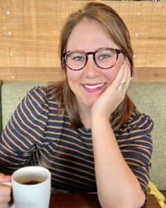 Smiling woman wearing glasses seated at a table holding a coffee mug, with a wooden wall background.