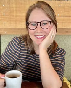 Smiling woman wearing glasses seated at a table holding a coffee mug, with a wooden wall background.