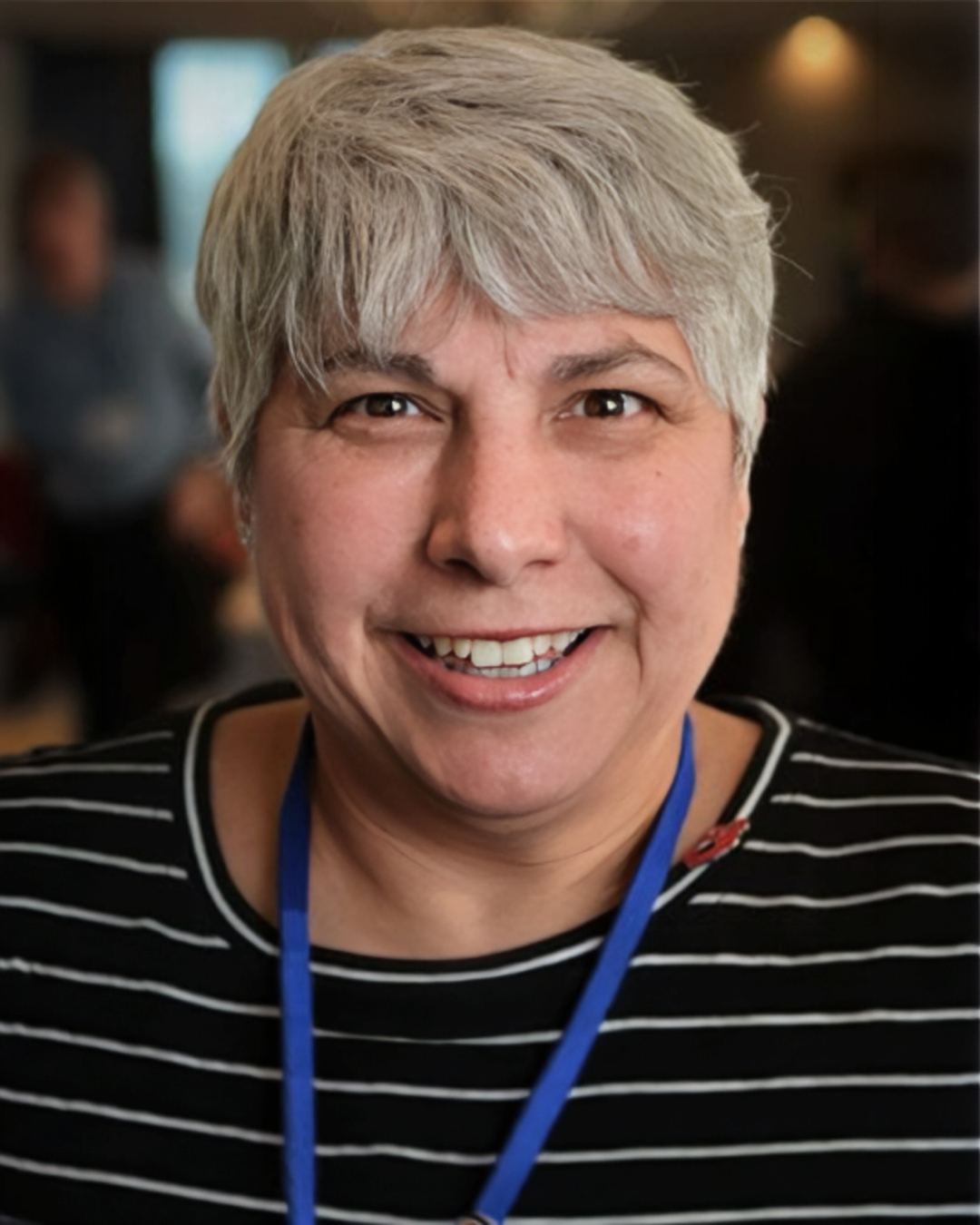 Smiling woman with short gray hair wearing a striped top and blue lanyard, photographed indoors with a softly blurred background.