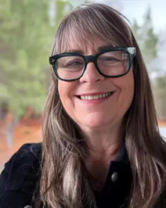 Smiling woman with long brown hair and glasses, photographed outdoors with trees softly blurred in the background.