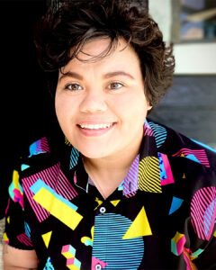 Smiling person with short dark hair wearing a colorful geometric-patterned button-up shirt, seated indoors with soft natural light in the background.