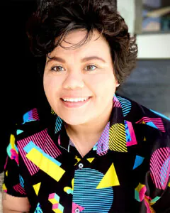 Smiling person with short dark hair wearing a colorful geometric-patterned button-up shirt, seated indoors with soft natural light in the background.