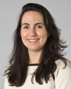 Professional headshot of a smiling woman with long dark hair wearing a light-colored top, photographed against a neutral gray background.