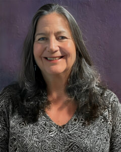 Portrait headshot of a woman with long dark hair wearing a patterned blouse, smiling against a purple background.