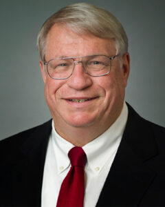 Formal headshot of a man wearing glasses, a black suit jacket, white shirt, and red tie.