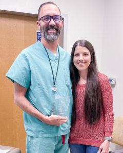 A healthcare professional in scrubs stands beside a smiling woman as they hold an award plaque together inside a clinical setting.