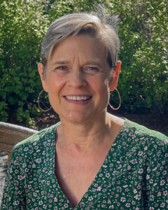 Outdoor portrait of a woman with short gray hair wearing a green floral dress, seated on a bench with greenery behind her.