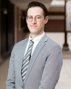 Professional headshot of a man wearing glasses, a gray suit, and striped tie, standing in an indoor setting.