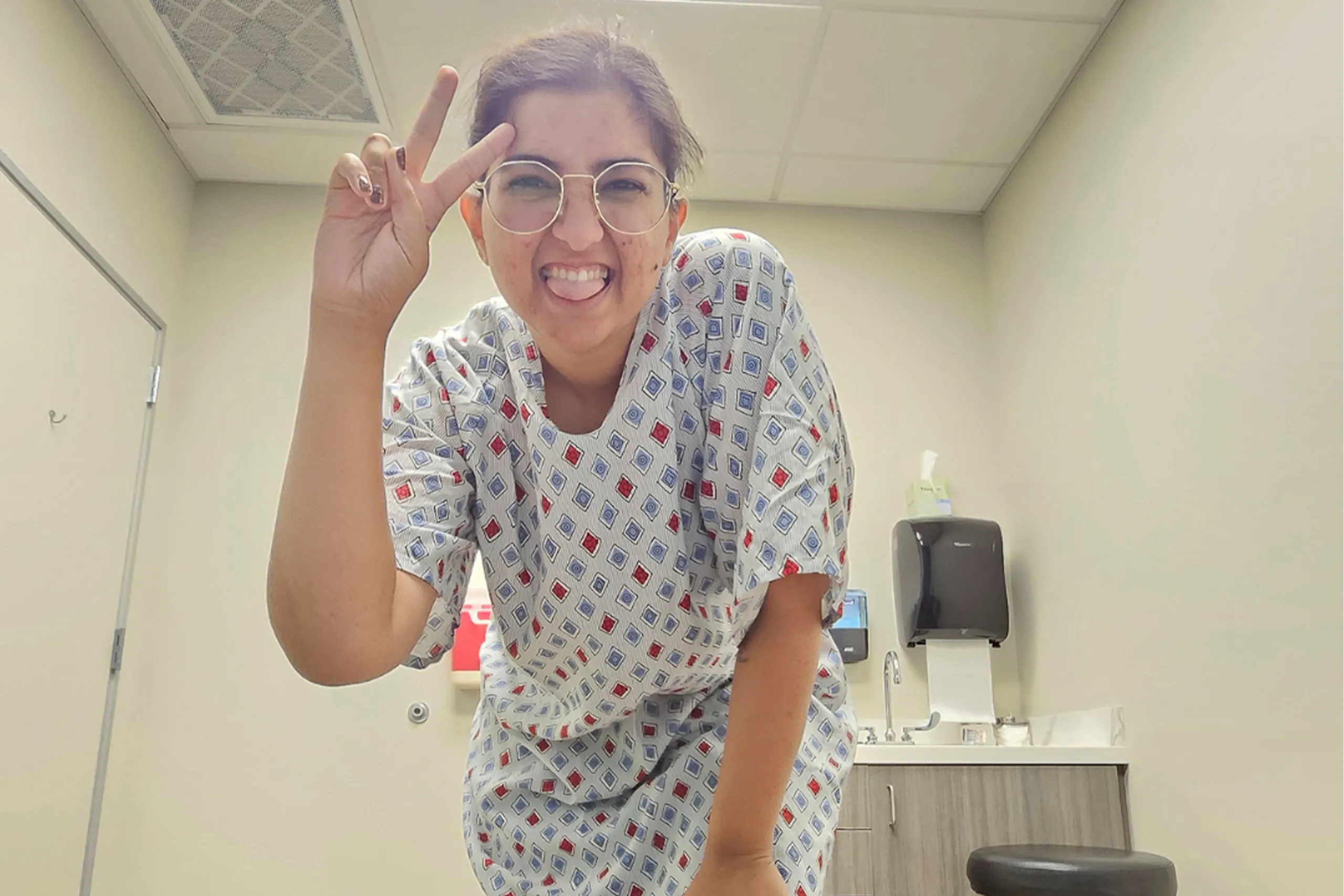 Smiling person wearing a hospital gown makes a peace sign toward the camera in a medical exam room.