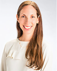 Professional headshot of a smiling woman with long brown hair wearing a light-colored blouse against a neutral background.