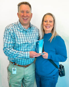 A man and woman smiling while holding an award together, standing against a plain indoor background.