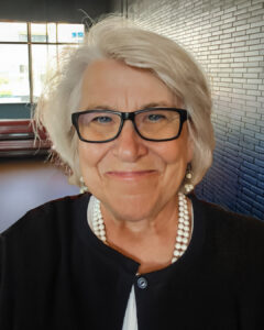 Portrait of a smiling woman with short white hair and black glasses wearing a black jacket and pearl necklace, photographed indoors with a brick wall and windows in the background.