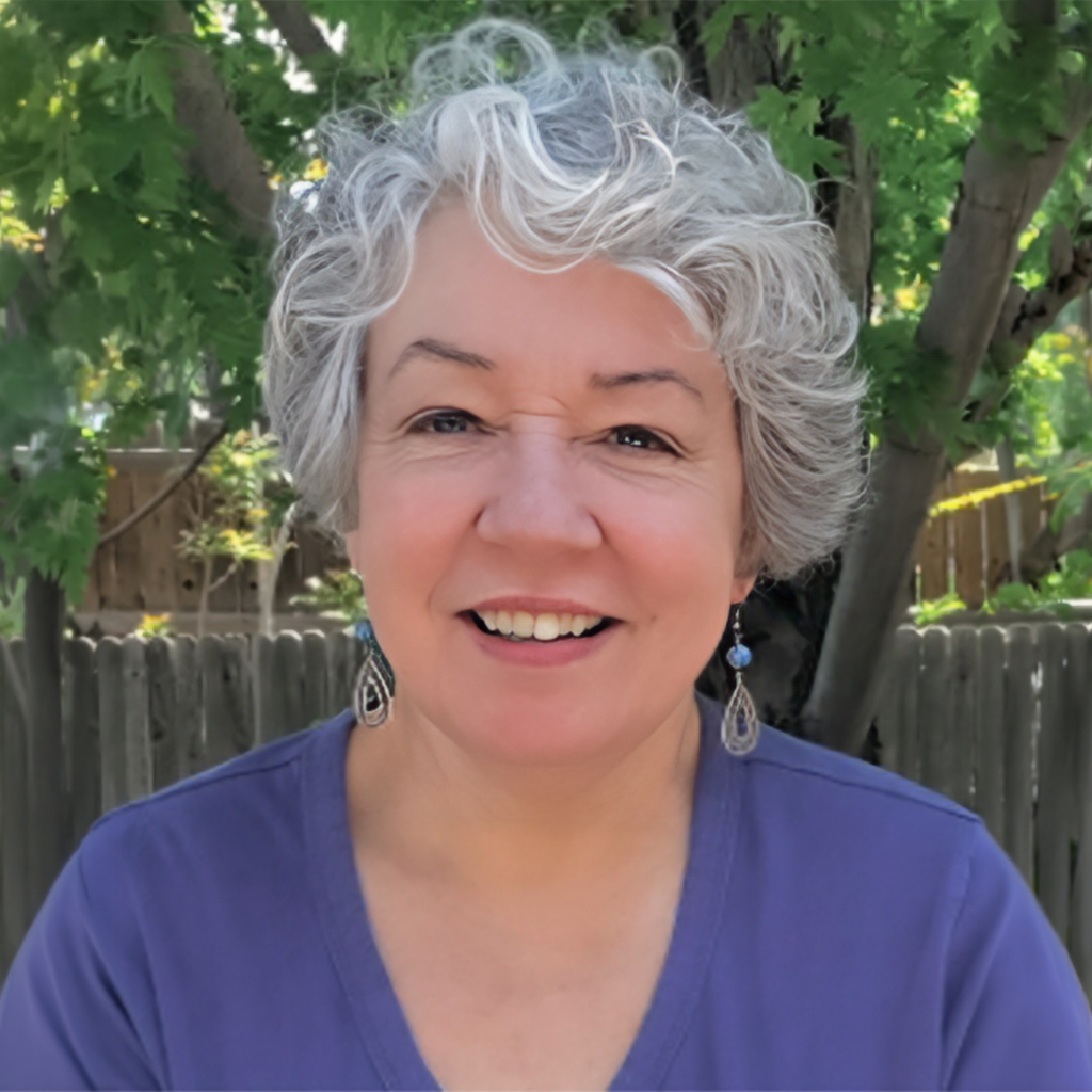 Smiling woman with short curly gray hair wearing a purple top, standing outdoors in front of trees and a wooden fence.