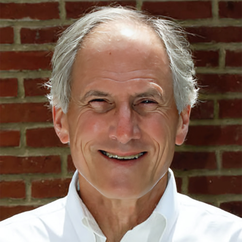 Smiling older man with gray hair wearing a white shirt, standing in front of a brick wall.