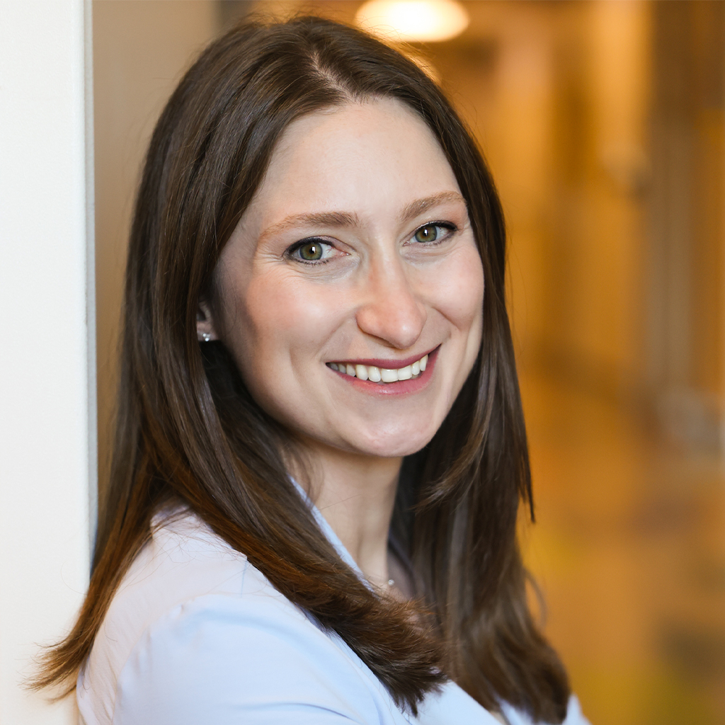 Smiling woman with long brown hair in a light blouse, photographed indoors with a softly blurred background.