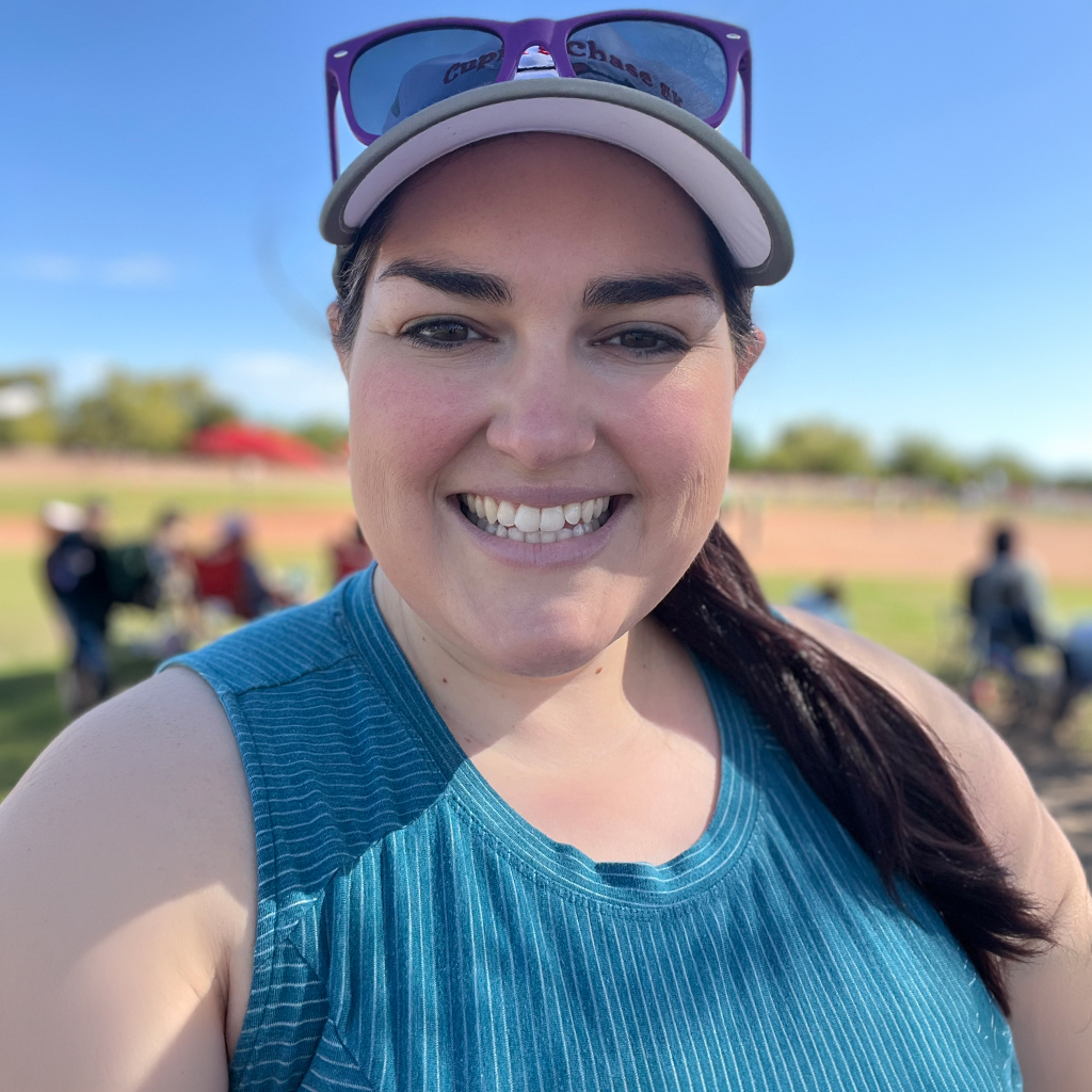 Smiling woman wearing a baseball cap and sunglasses on her head, outdoors at a field on a sunny day.