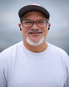 Casual outdoor portrait of a smiling man with glasses and a gray beard wearing a baseball cap, white sweatshirt, and wireless earbuds, posed against a softly blurred gray background.
